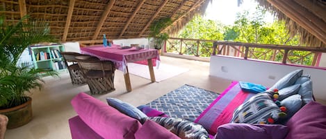 A room with a thatched roof, a sofa with cushions, a table with a pink tablecloth, and a view of greenery outside.