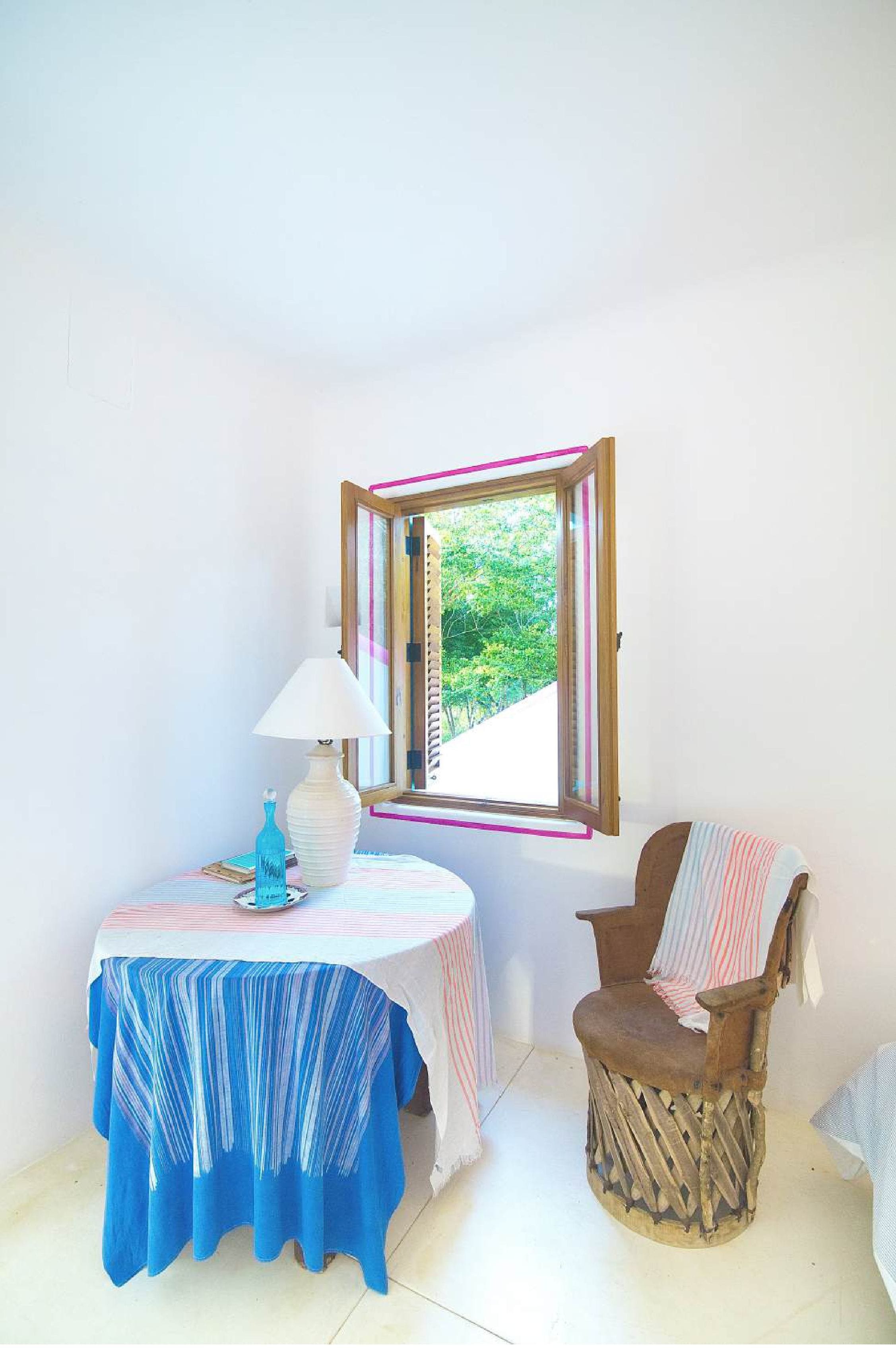 A small room with a table and chair, a window with a view of greenery, and a lamp.