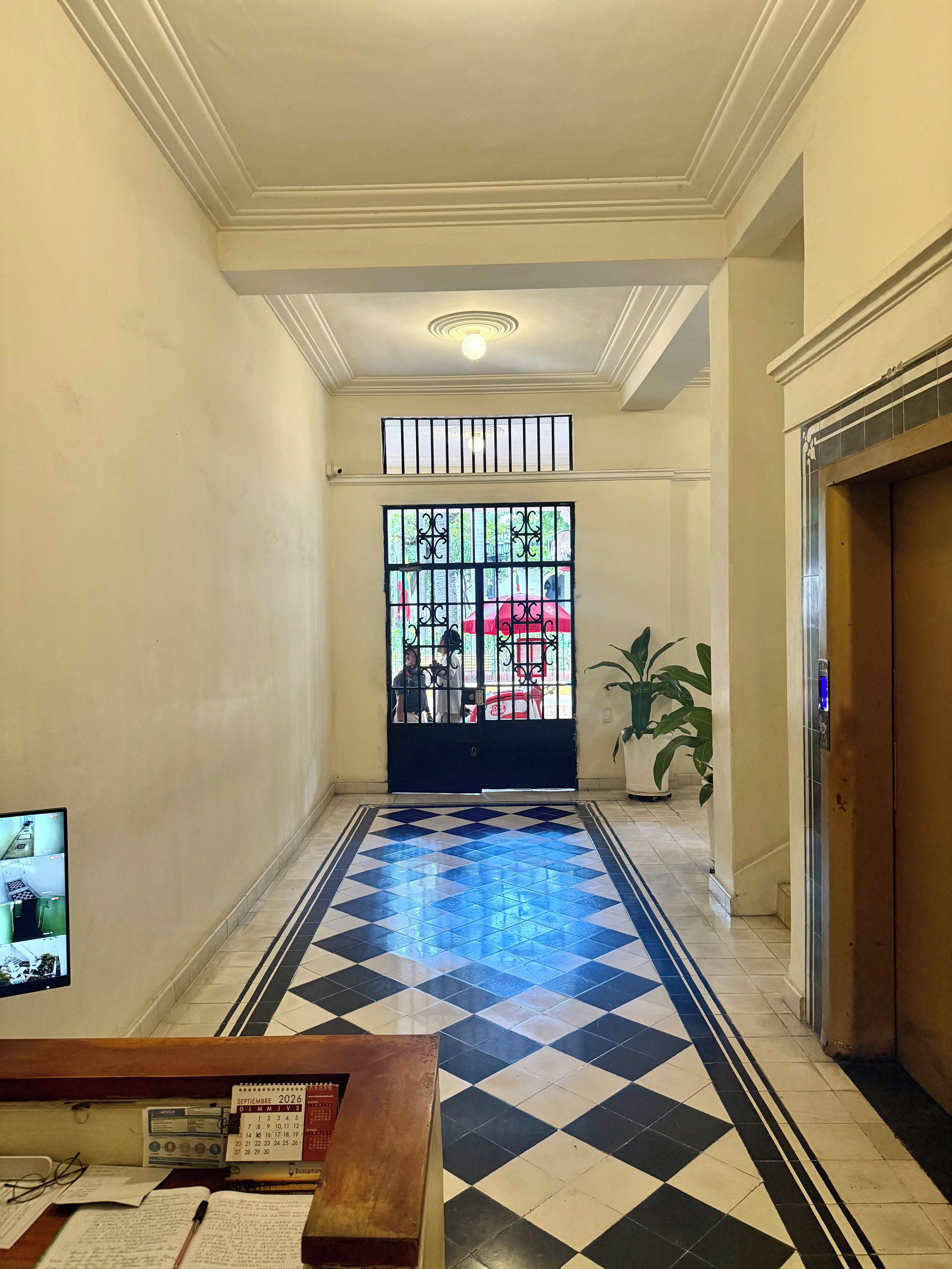 A hotel lobby with a check-in counter, a tiled floor, a large window with bars, and a potted plant.