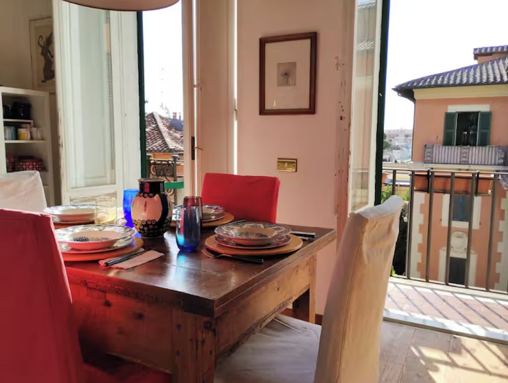 Uma área de refeições com mesa de madeira, cadeiras vermelhas e vista para edifícios por uma grade de varanda.