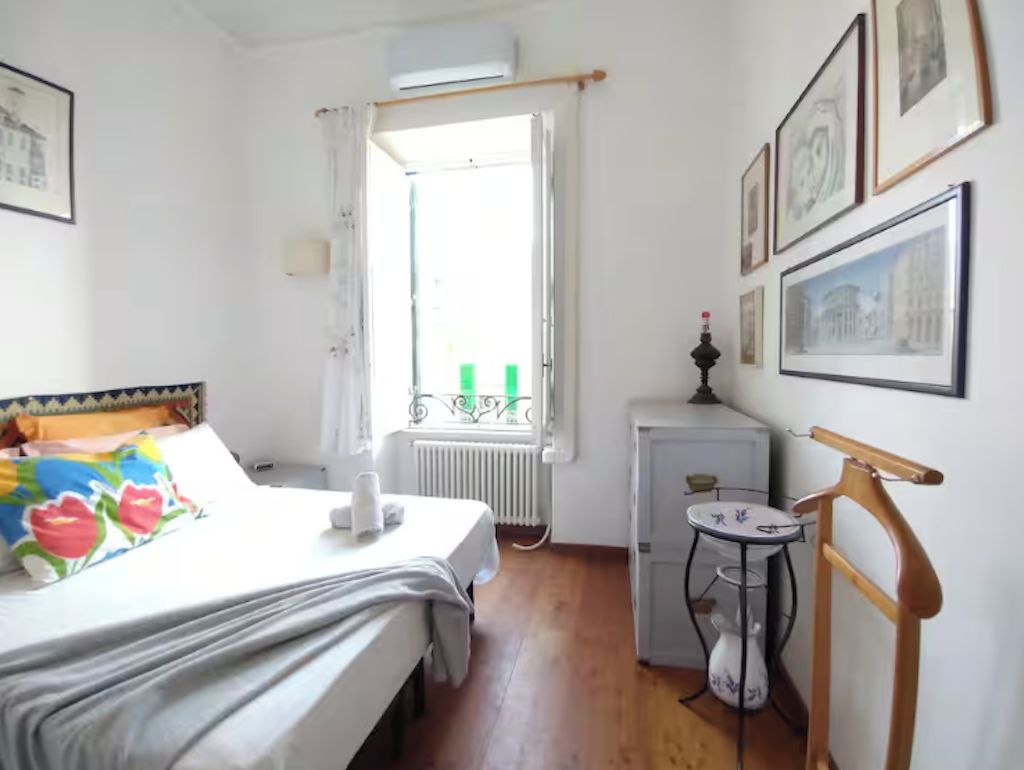 A bedroom with a bed, a bedside table, a radiator, a window with curtains, and framed pictures on the wall.