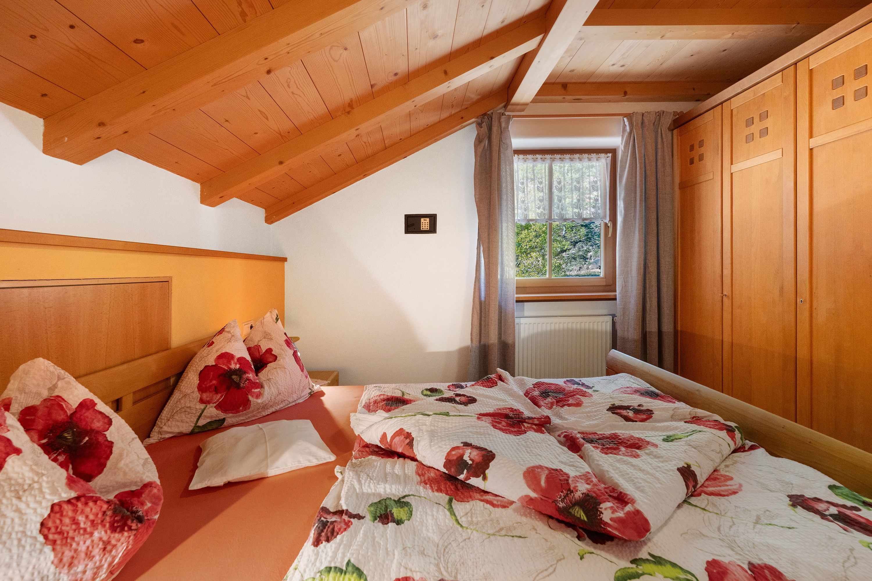 Une chambre avec un plafond en bois, un lit avec une literie à motifs floraux, une fenêtre avec des rideaux et un radiateur.