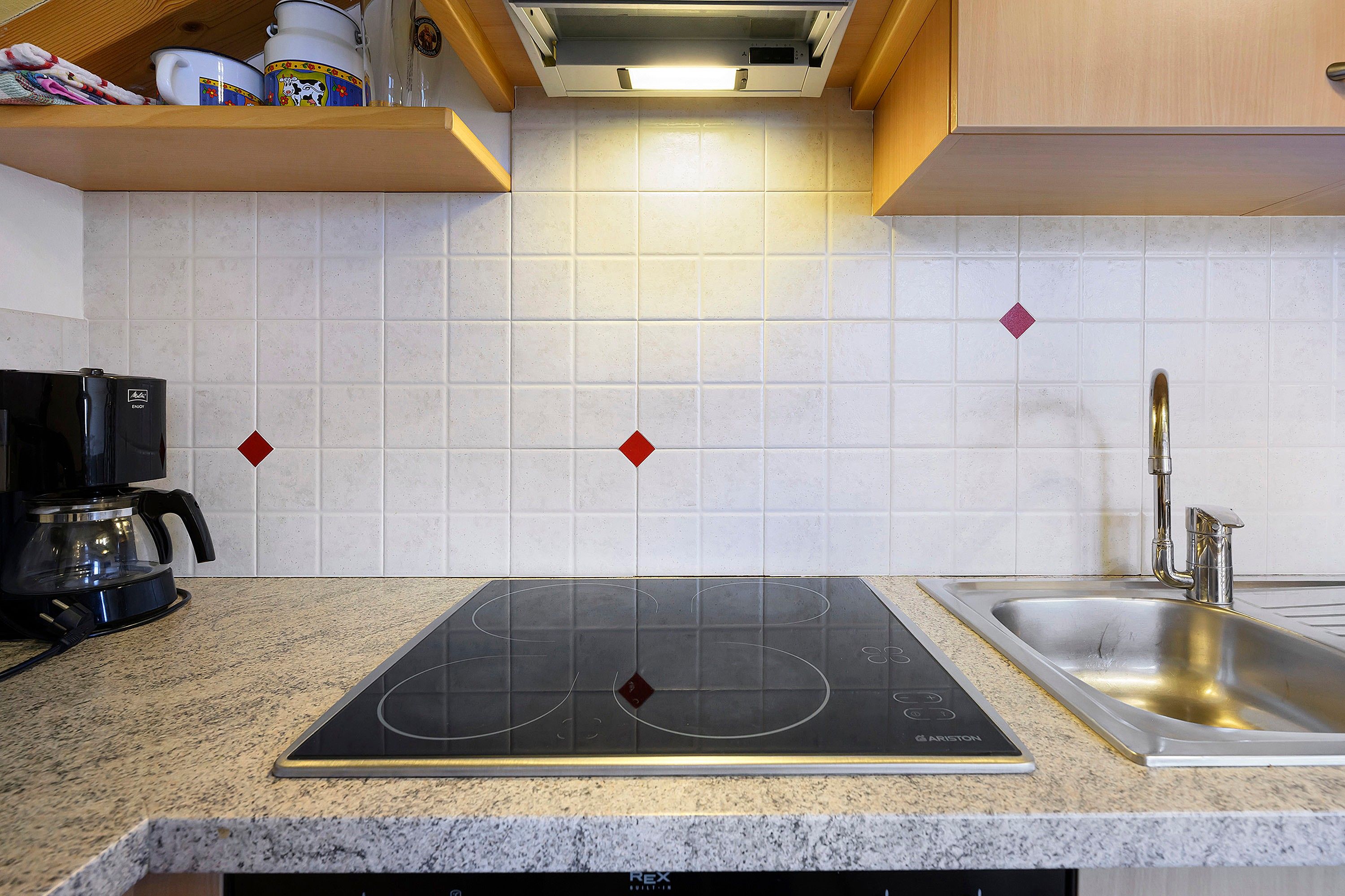 Una cucina con piano cottura a induzione nero, lavello in acciaio inossidabile e pareti rivestite di piastrelle bianche.