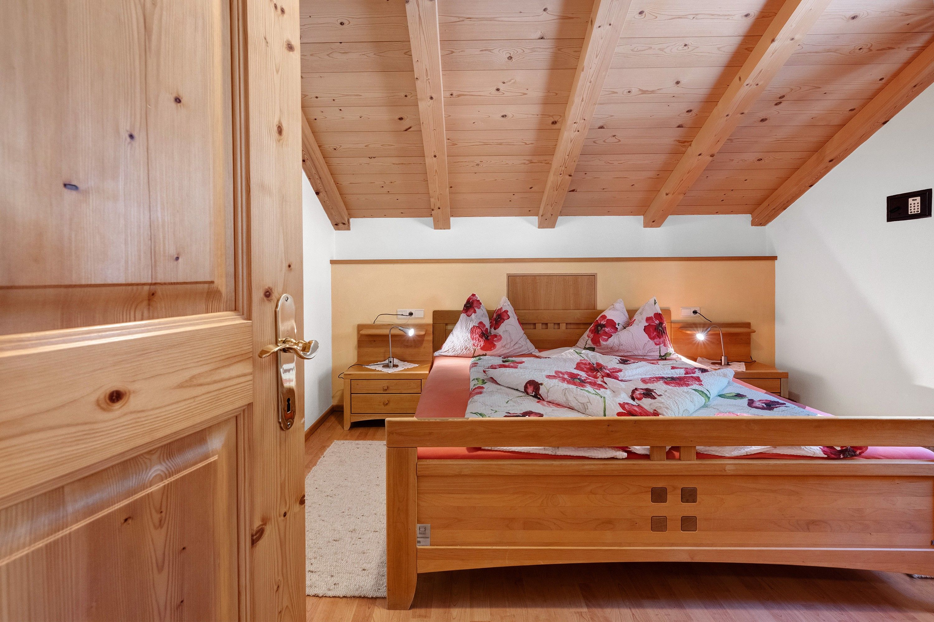 Una camera da letto con soffitto in legno, un letto con biancheria da letto floreale, un comodino con una lampada e una sveglia fissata al muro.