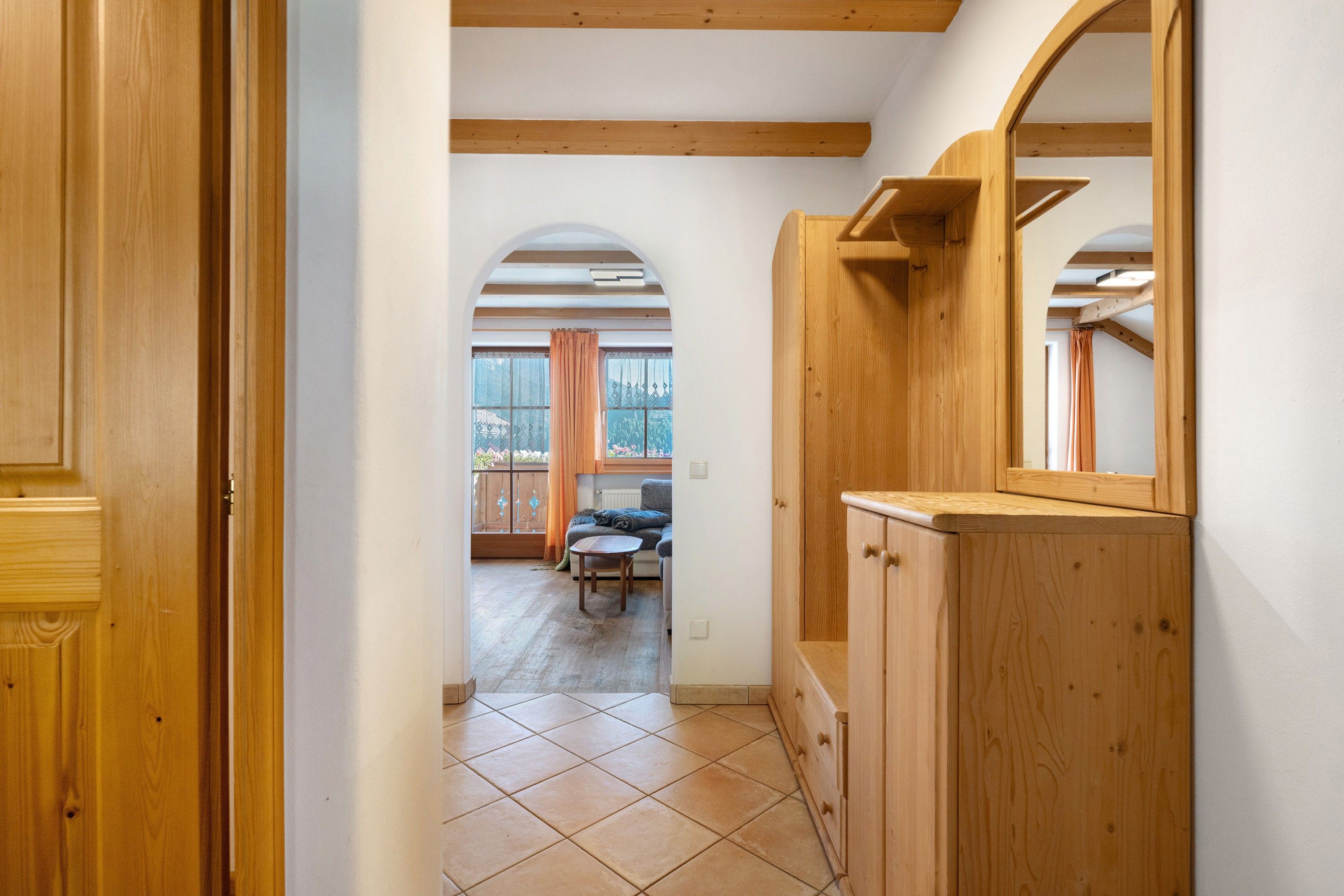 Un couloir avec des portes en bois, un sol carrelé et une vue sur une pièce où se trouvent un lit et une fenêtre.