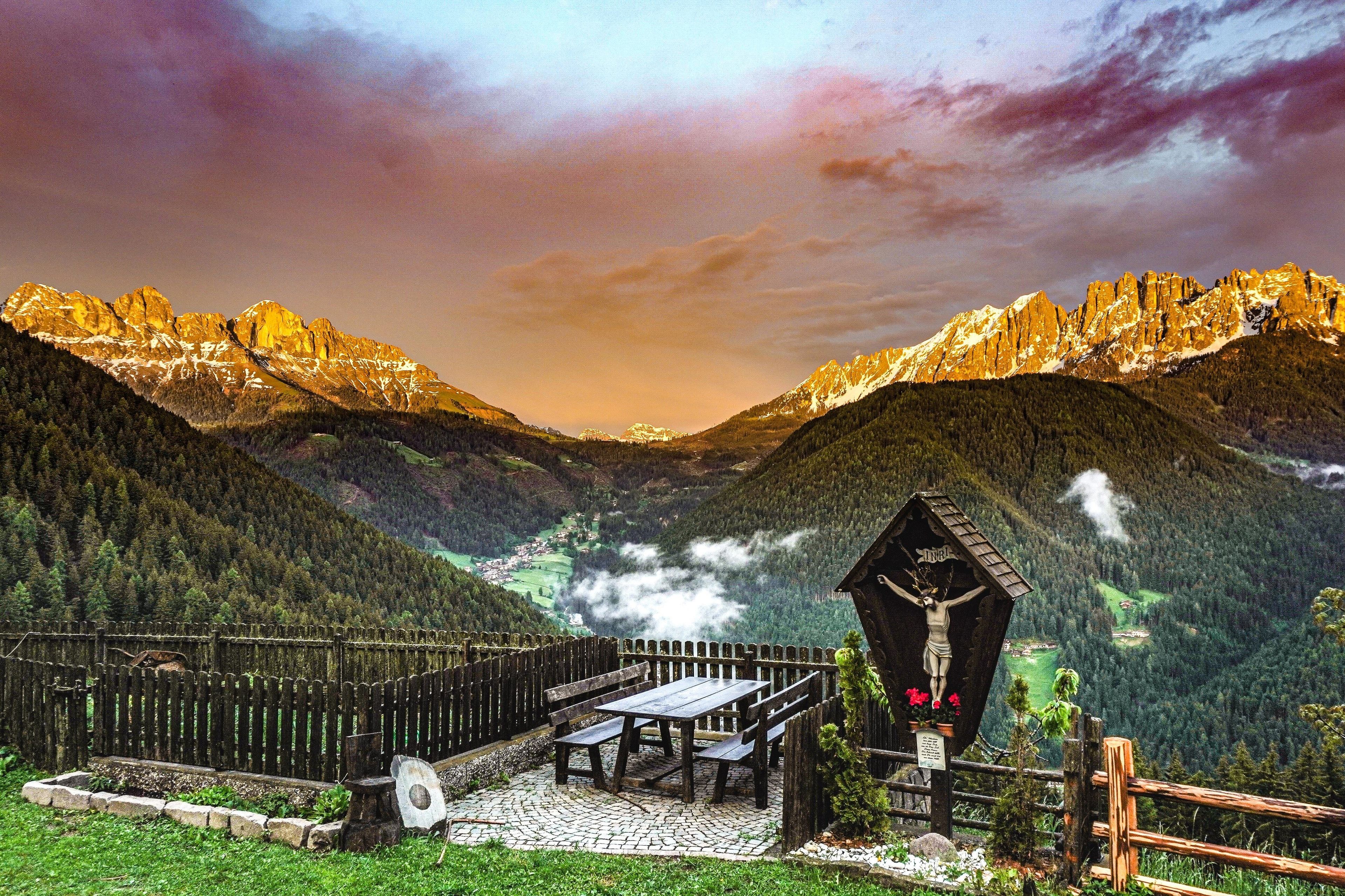 A mountain landscape with a wooden structure featuring a crucifix, a picnic table, and benches.