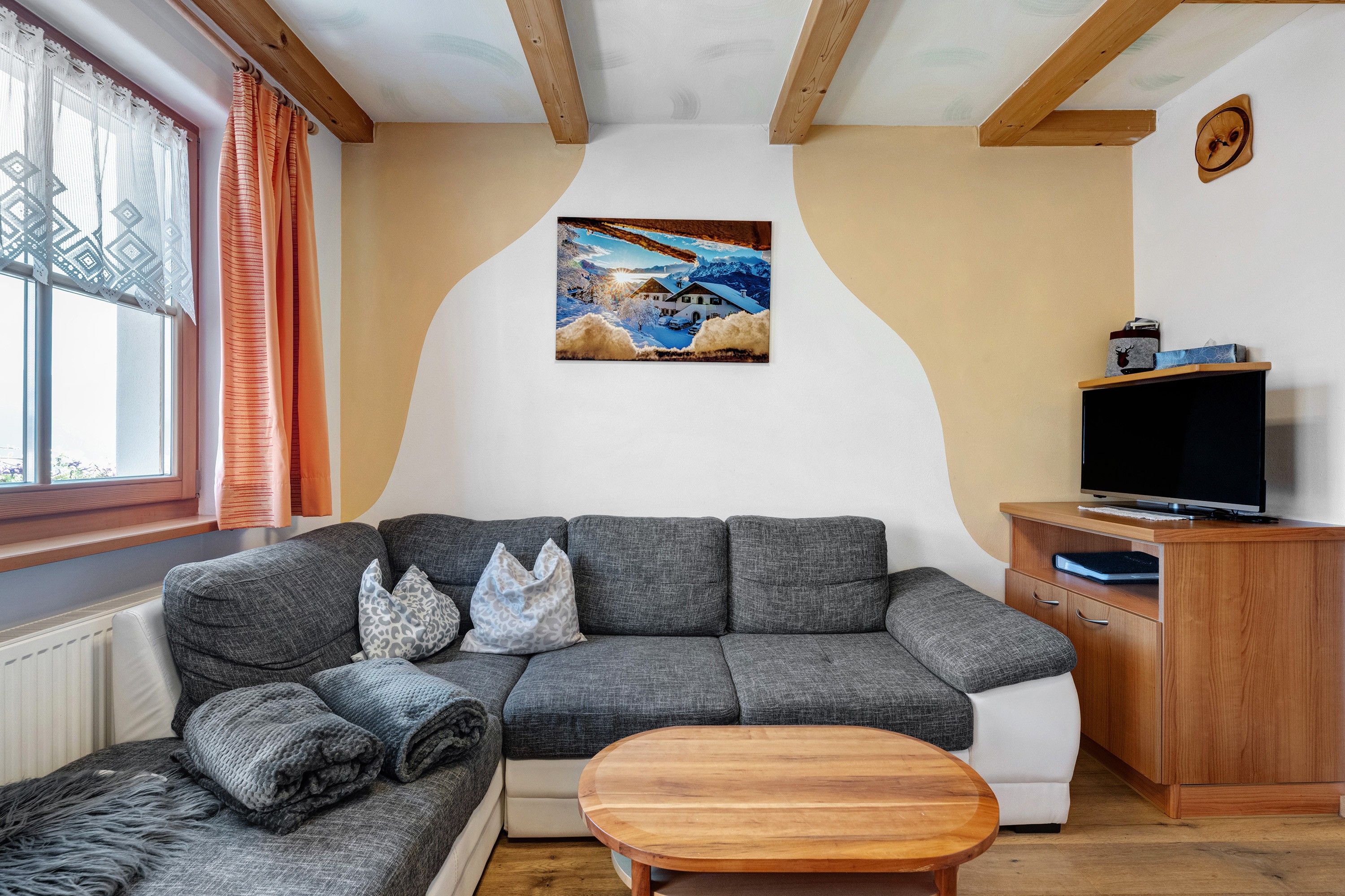 A living room with a grey sectional sofa, a wooden coffee table, a TV on a wooden cabinet, and a painting of a snowy landscape on the wall.