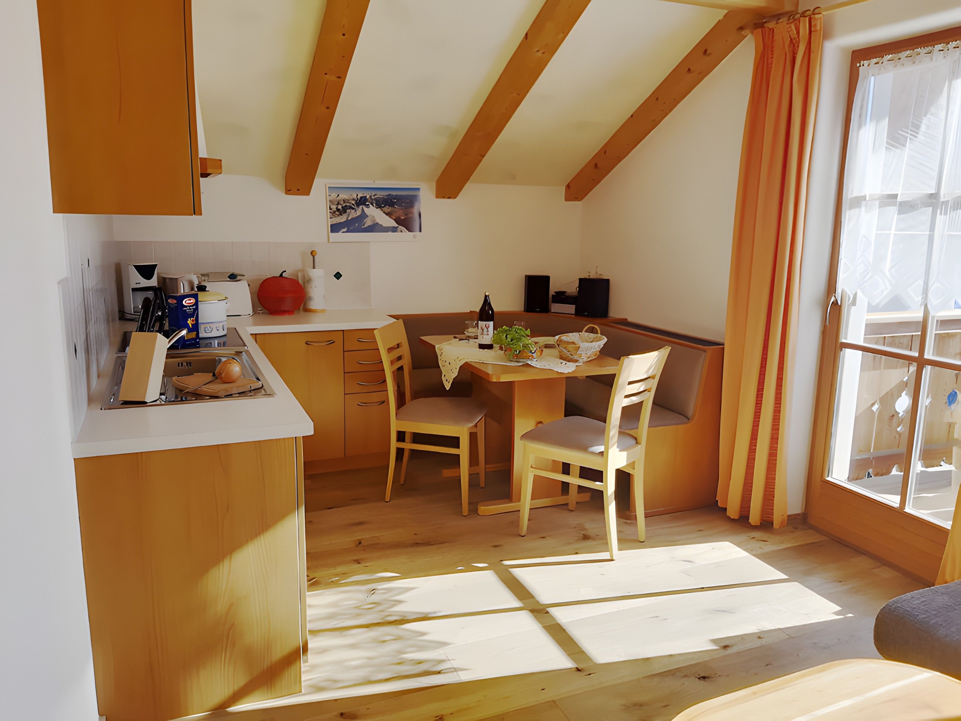 A compact kitchen with wooden cabinets, a dining table with chairs, and a view through a window.