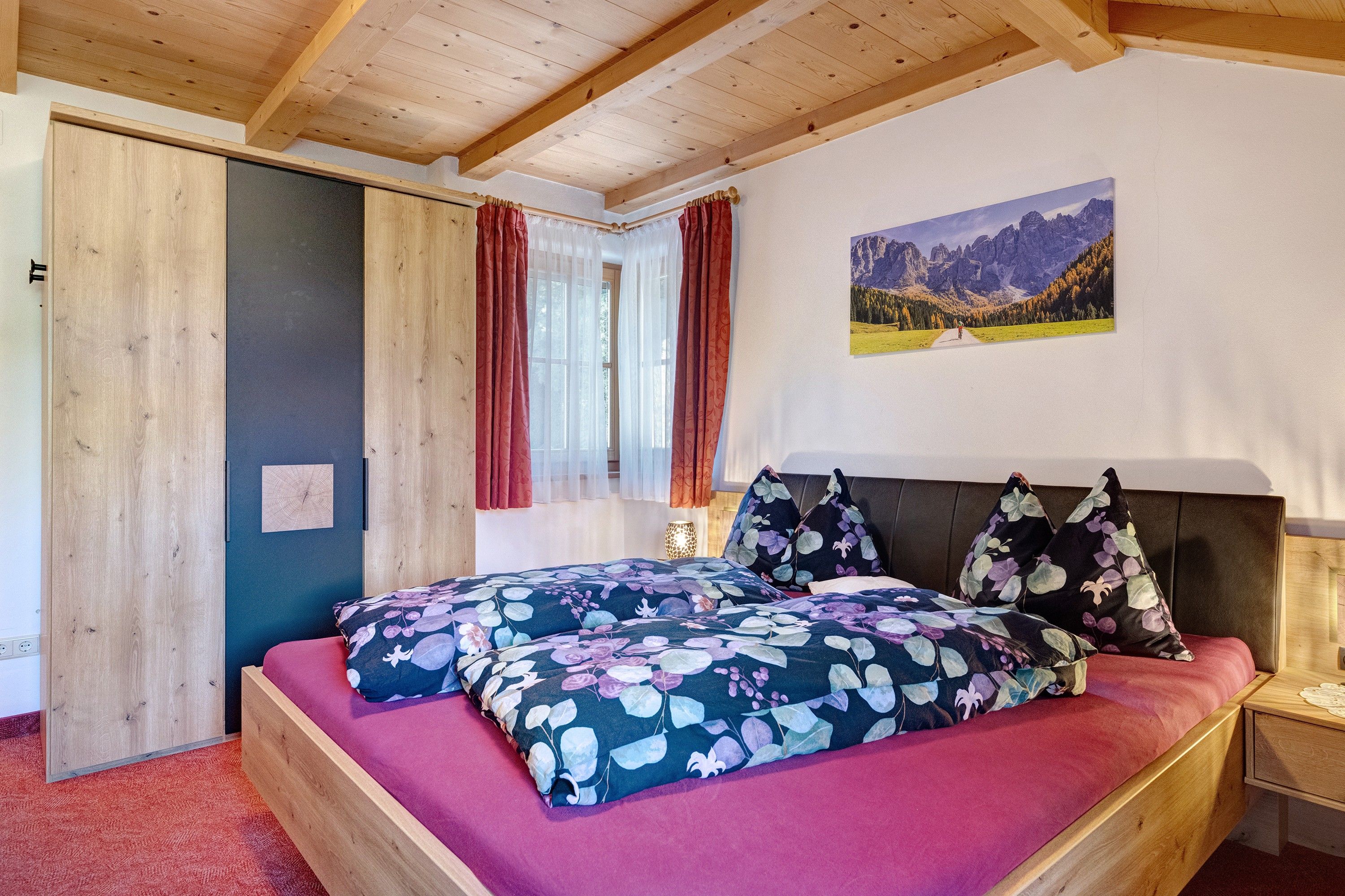 Une chambre avec un plafond en bois, un lit recouvert d’une literie à motifs floraux, une table de chevet et un tableau représentant un paysage de montagne accroché au mur.