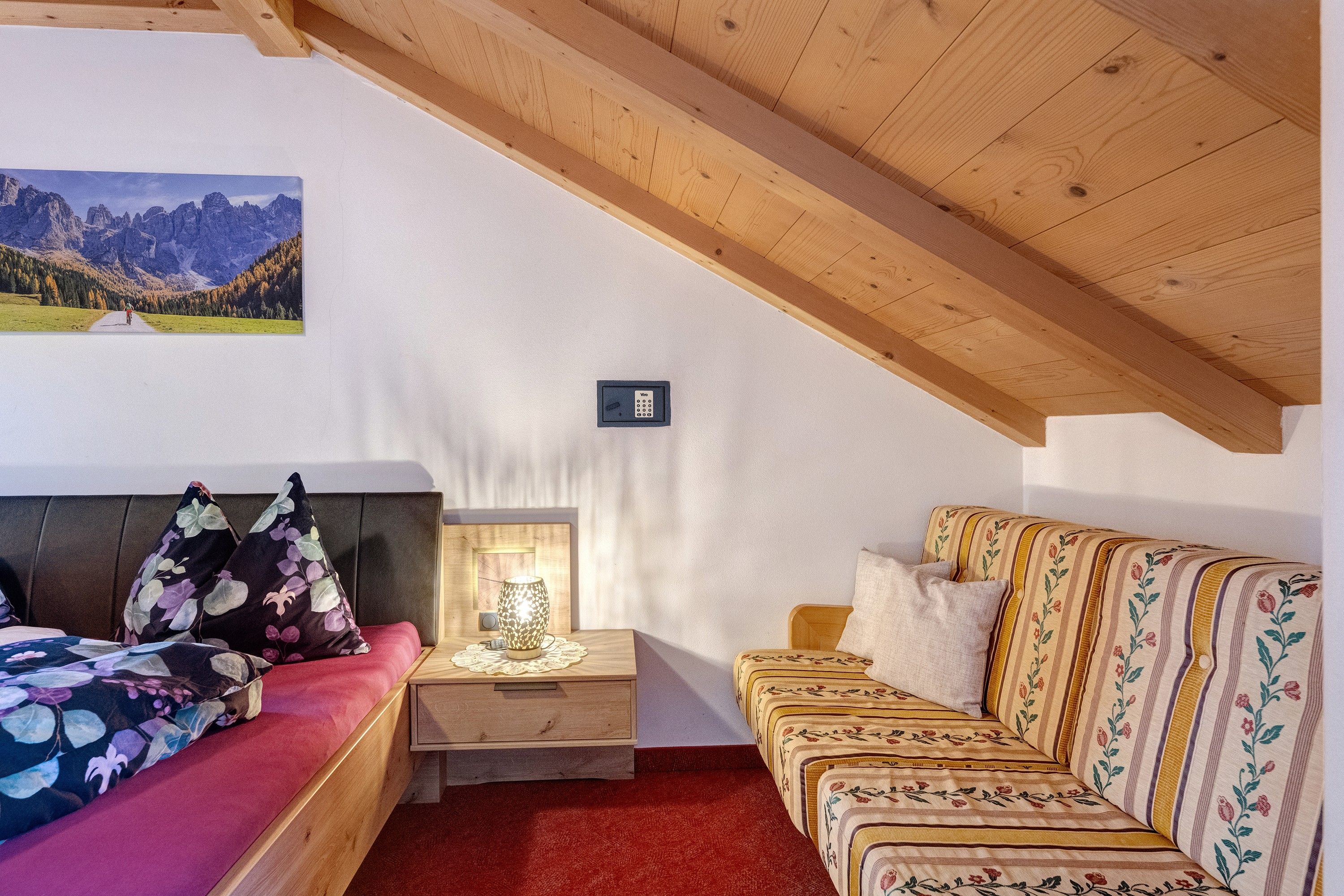 Una stanza con un letto, un divano, un comodino con una lampada, un quadro appeso al muro e un soffitto in legno.