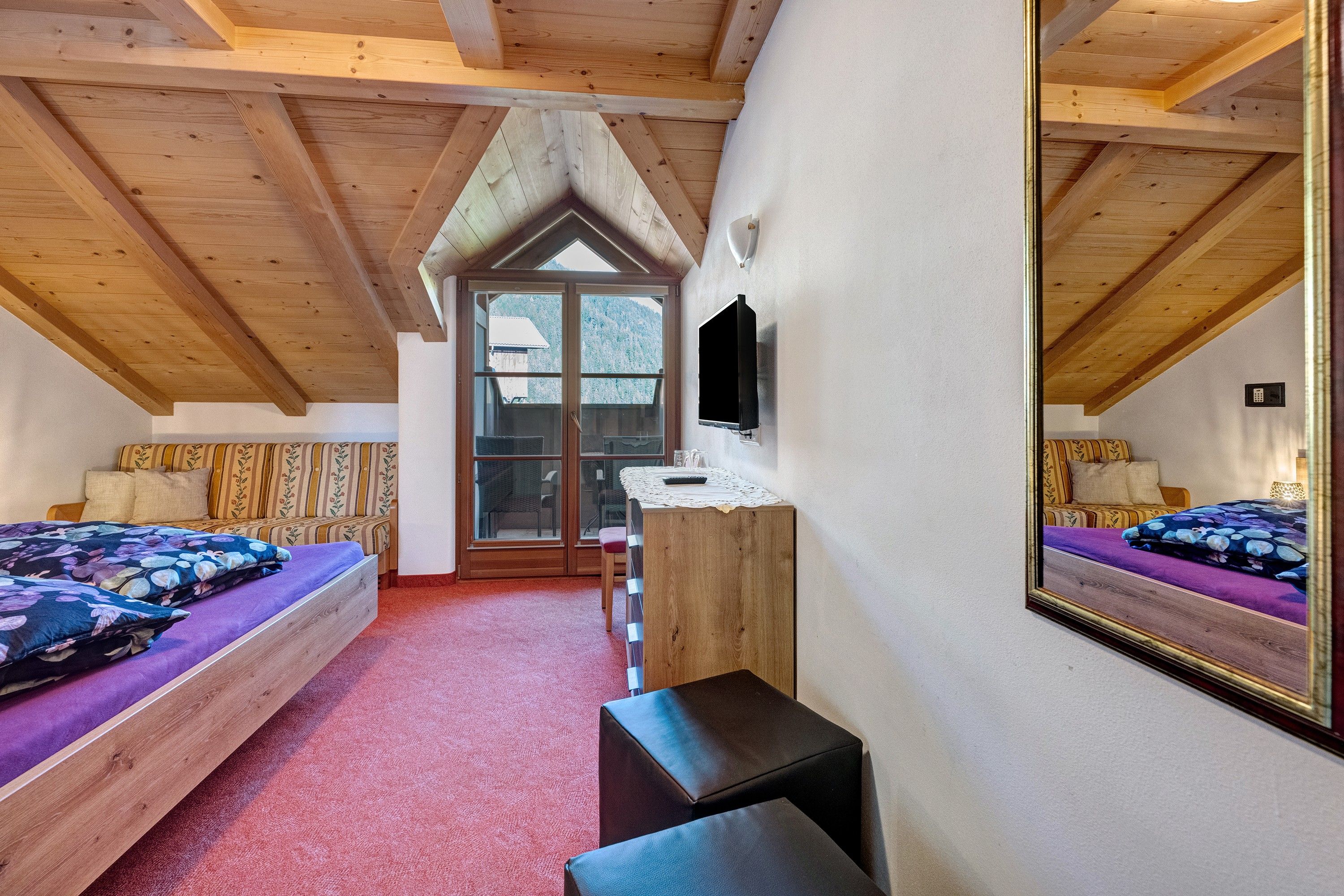 Une chambre avec un plafond en bois, un lit recouvert d’une literie à motifs floraux, un banc et un téléviseur.