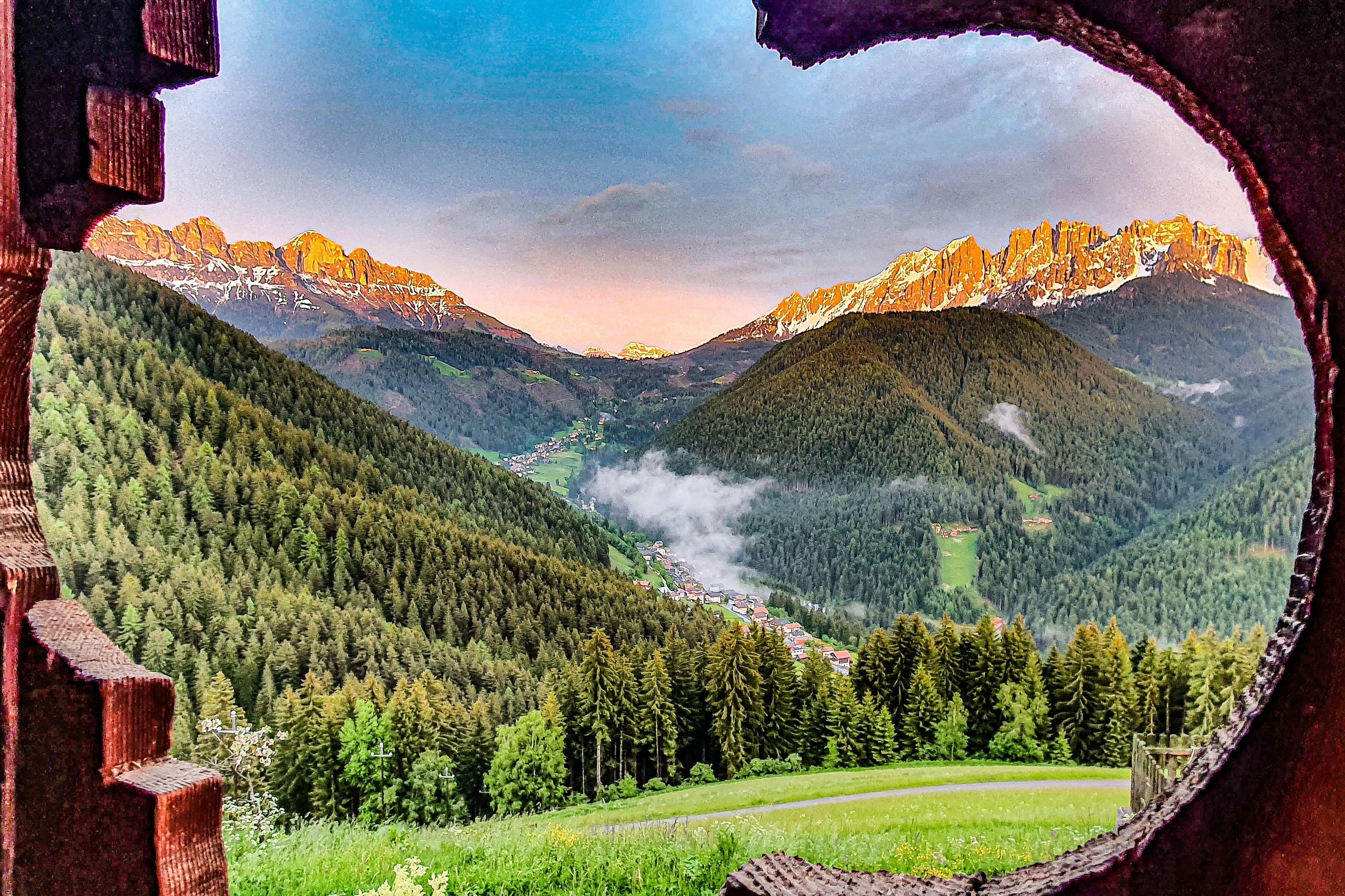 A panoramic view of a mountainous landscape through a circular window, featuring dense forests, a valley, and distant peaks.