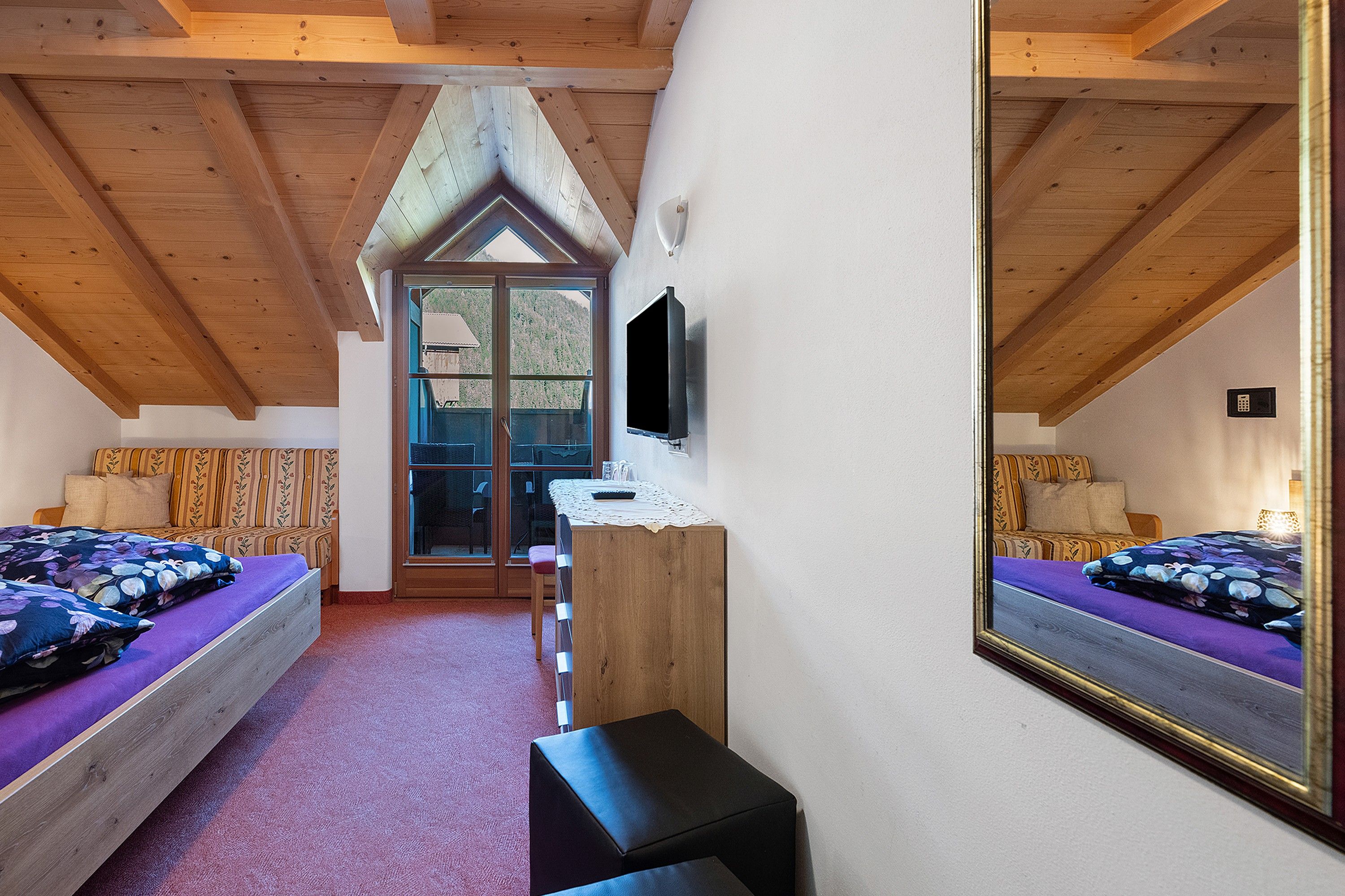 A bedroom with a sloped ceiling, a bed with a floral bedspread, a small table with a television, and a window with a view of trees.