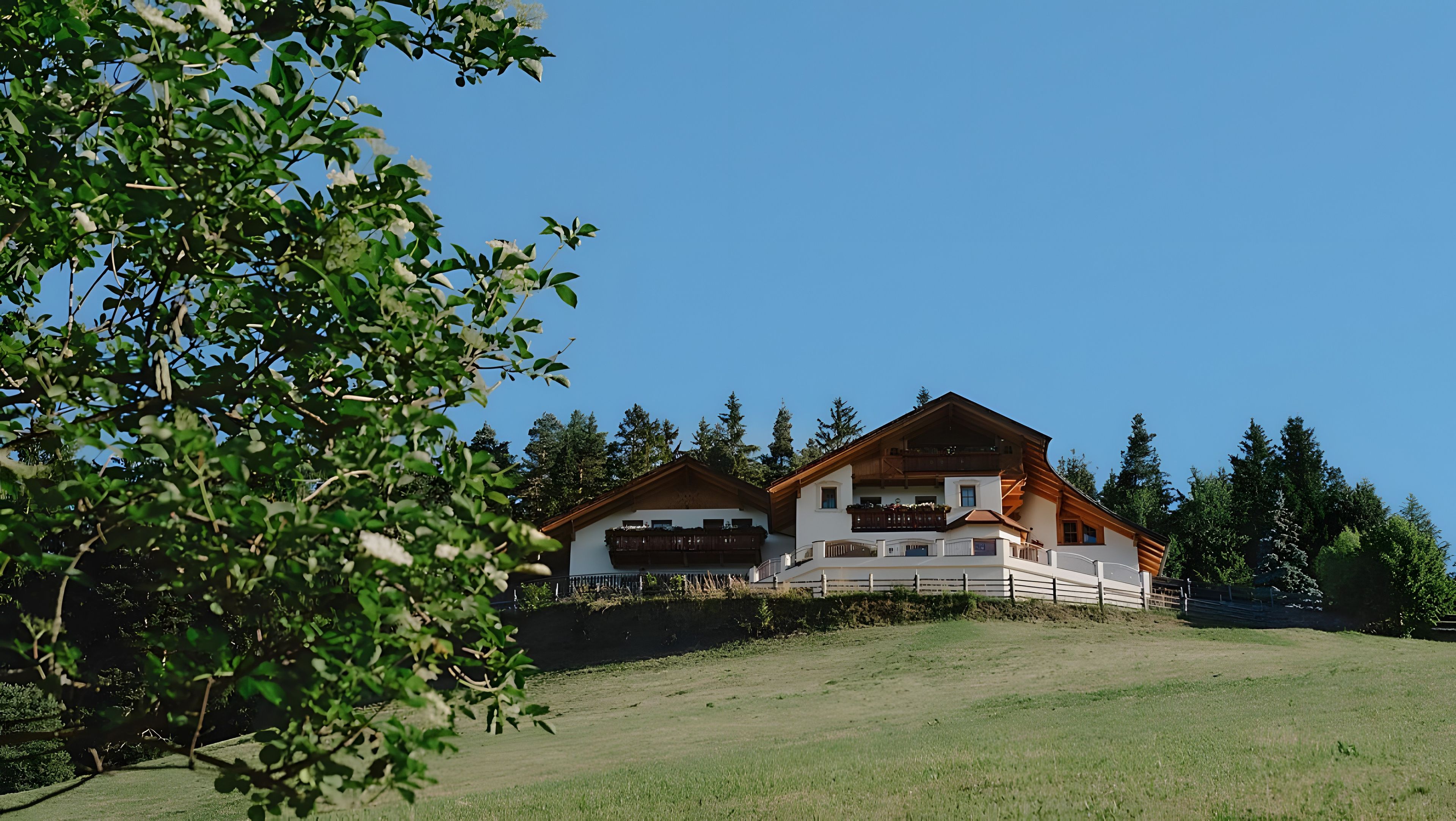 Un chalet à deux étages doté d’un balcon et d’une grande fenêtre, entouré d’arbres et d’une colline verdoyante.