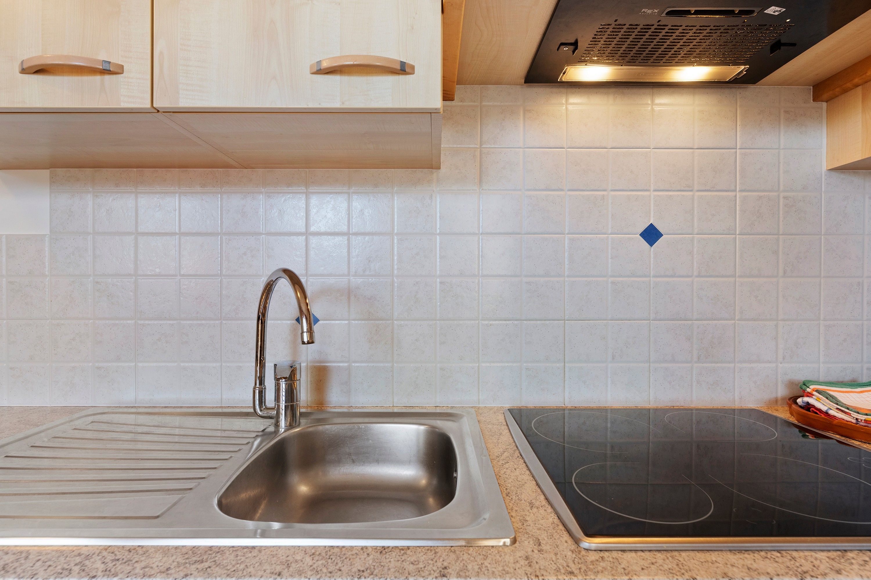 Une cuisine dotée d’un évier en acier inoxydable, d’une plaque de cuisson à induction noire et de murs recouverts de carreaux de couleur claire.