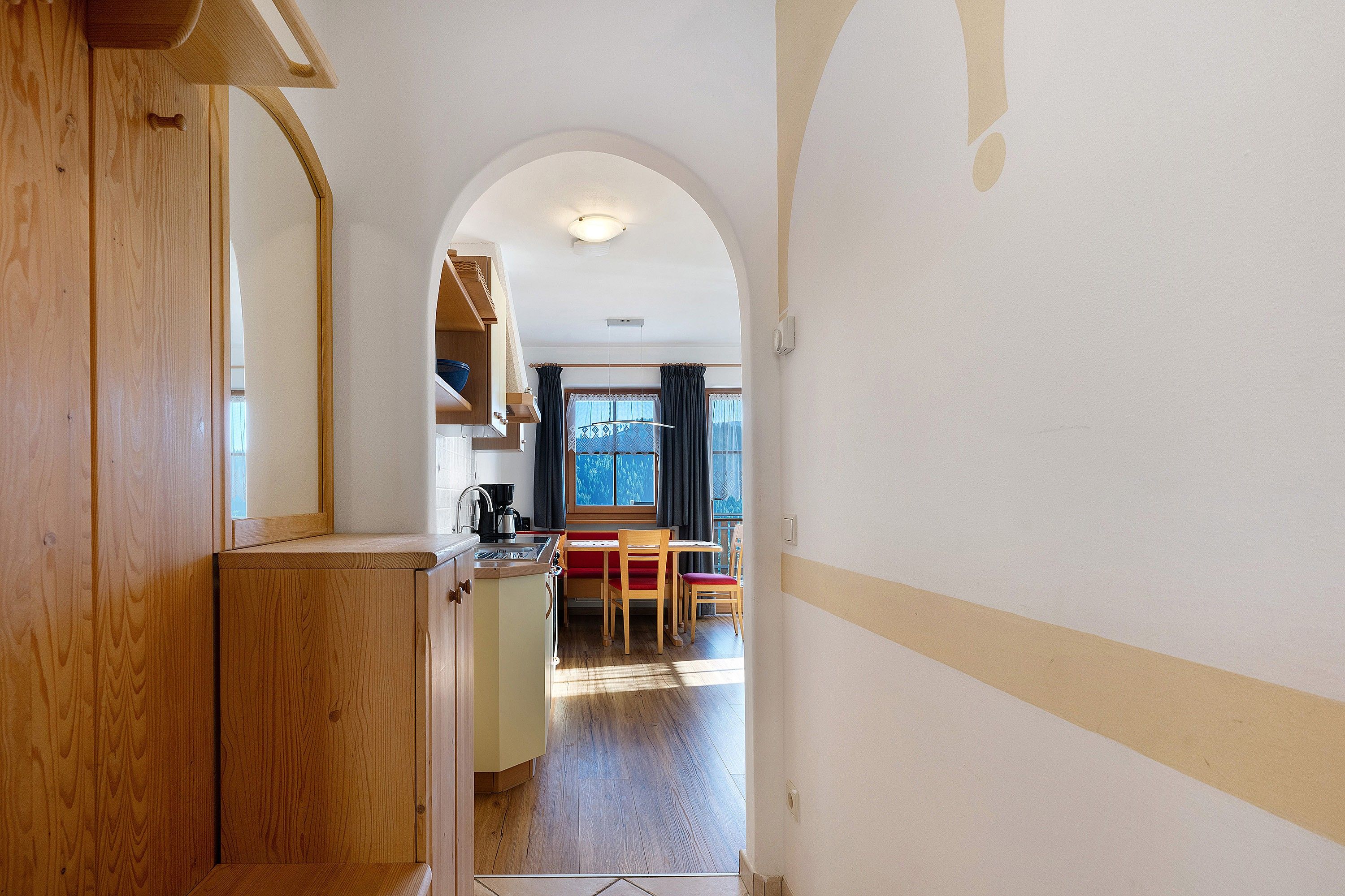 A narrow hallway leading to a dining area with a wooden table and chairs, a window with curtains, and a kitchen area with a sink and cabinets.