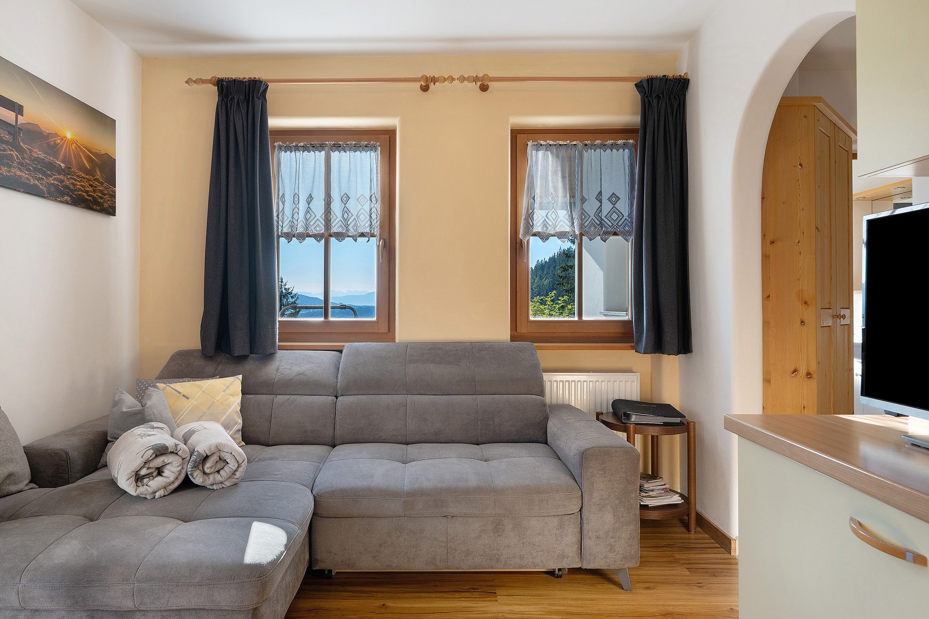 Un salon moderne avec un canapé gris, une petite table d’appoint et un téléviseur. Il y a deux fenêtres avec des rideaux, un radiateur et un meuble en bois.