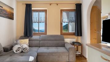 Un salon moderne avec un canapé gris, une petite table d’appoint et un téléviseur. Il y a deux fenêtres avec des rideaux, un radiateur et un meuble en bois.