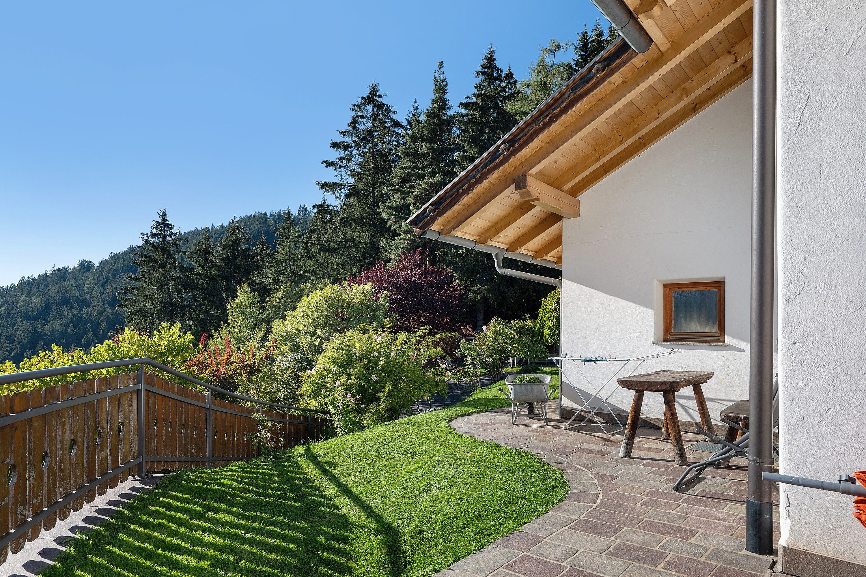 Une maison avec un escalier en bois menant à un balcon, un jardin avec une pelouse et diverses plantes, et une vue sur une colline boisée.