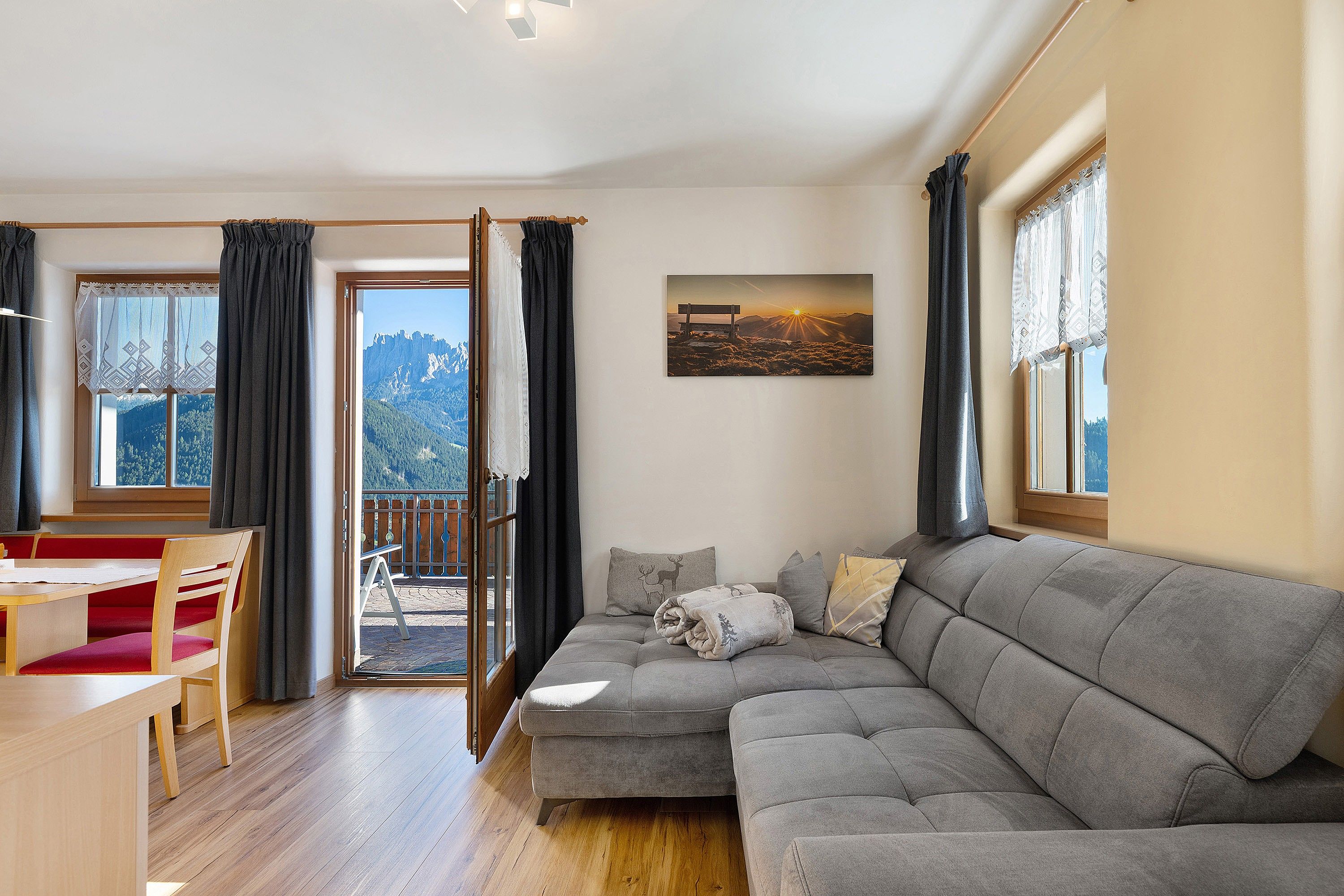 A cozy living room with a grey sofa, a wooden dining table with red chairs, and a balcony with a mountain view.