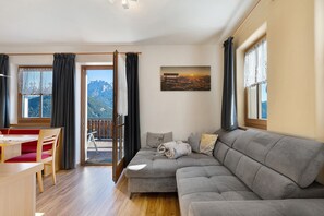 Un salón acogedor con un sofá gris, una mesa de comedor de madera con sillas rojas, y un balcón con vistas a la montaña.