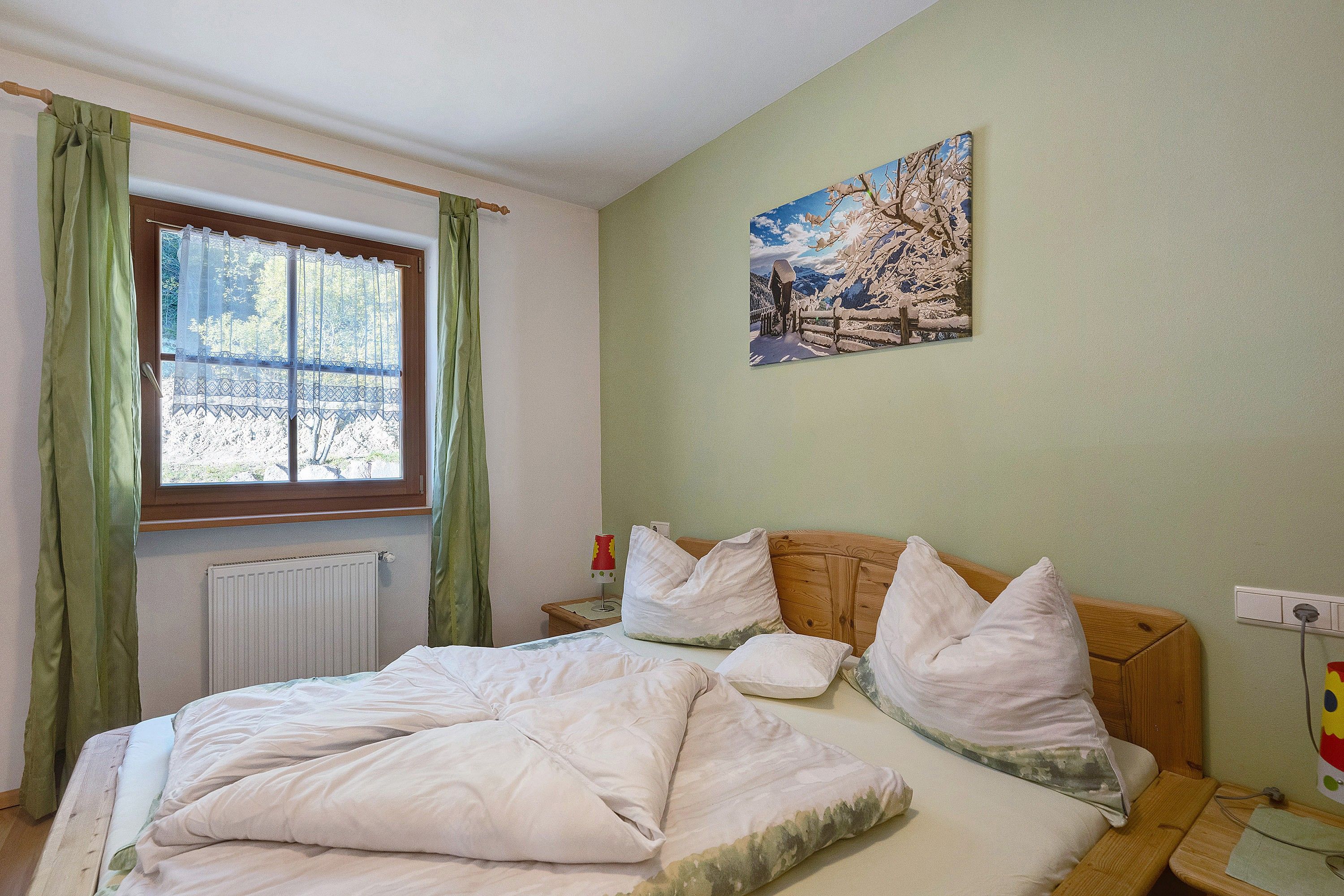 A bedroom with a bed, a window with curtains, a painting on the wall, and a bedside table with a lamp.