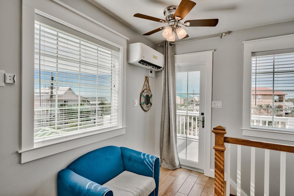 A room with a ceiling fan, large windows with blinds, a blue sofa, and a view of the outside.