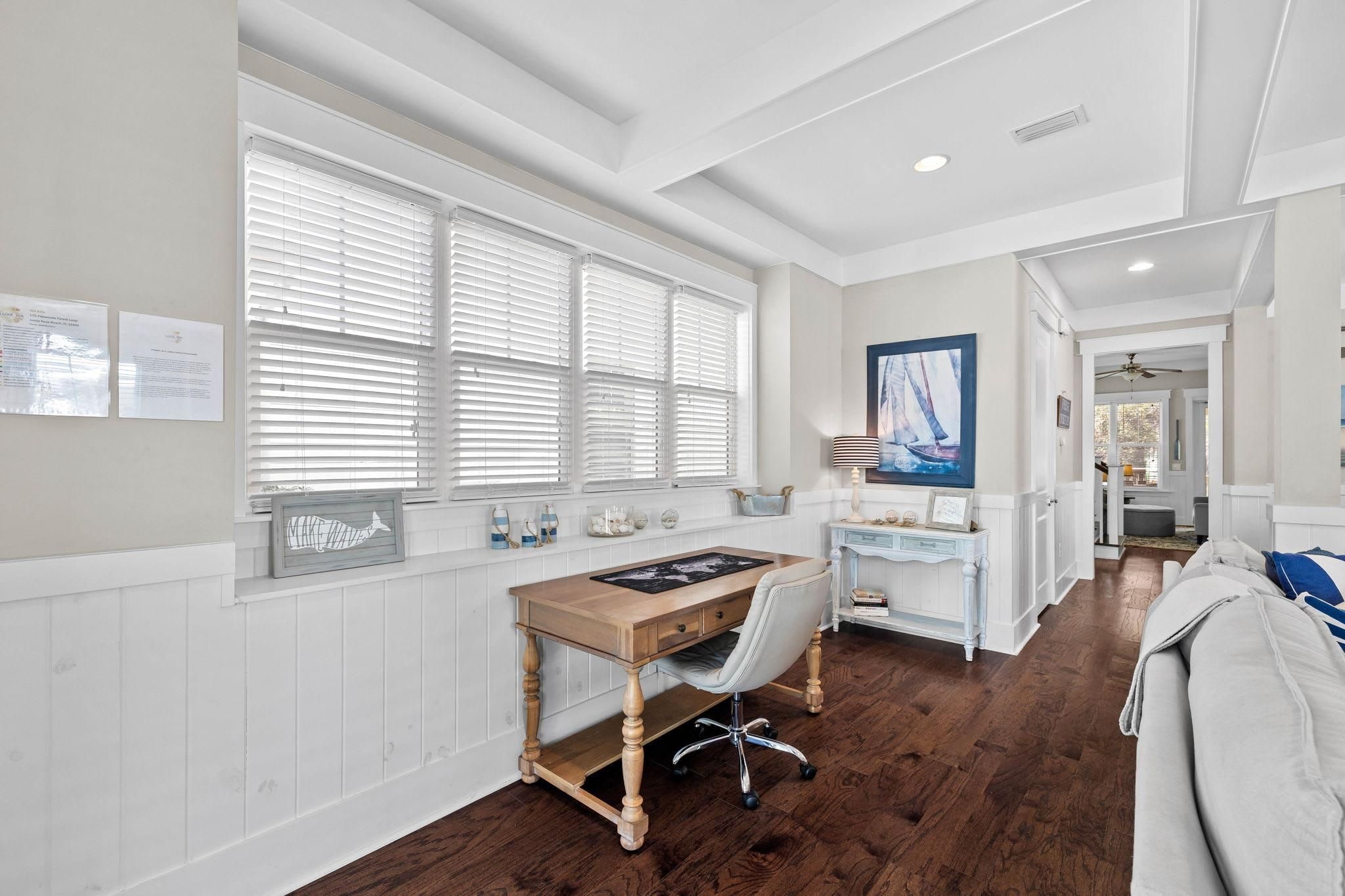 A home office with a wooden desk, white walls, and a view through large windows.