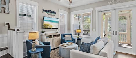 A well-lit living room with a ceiling fan, a television, and a view of the outdoors through large windows.