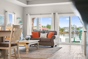 A cozy living room with a view of a body of water, a dining area with a table and chairs, and a large window with curtains. - Angler Dock House (Panama City Beach)