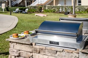 Een barbecue met een stenen onderstel, twee borden met burgers, en een gazon met stoelen en een gebouw op de achtergrond.