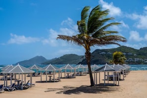 A beach with beach umbrellas and lounge chairs, a palm tree, and a mountain in the background. - Top-View Oceanside 2BR Villa, Beachfront Marriott St. Kitts – Dec 8–15, Sleeps 8 (Frigate Bay)