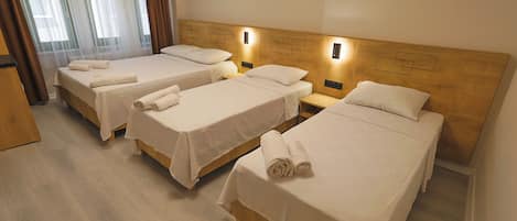A hotel room with three single beds, each with a neatly folded towel, wooden headboards, and bedside lamps.