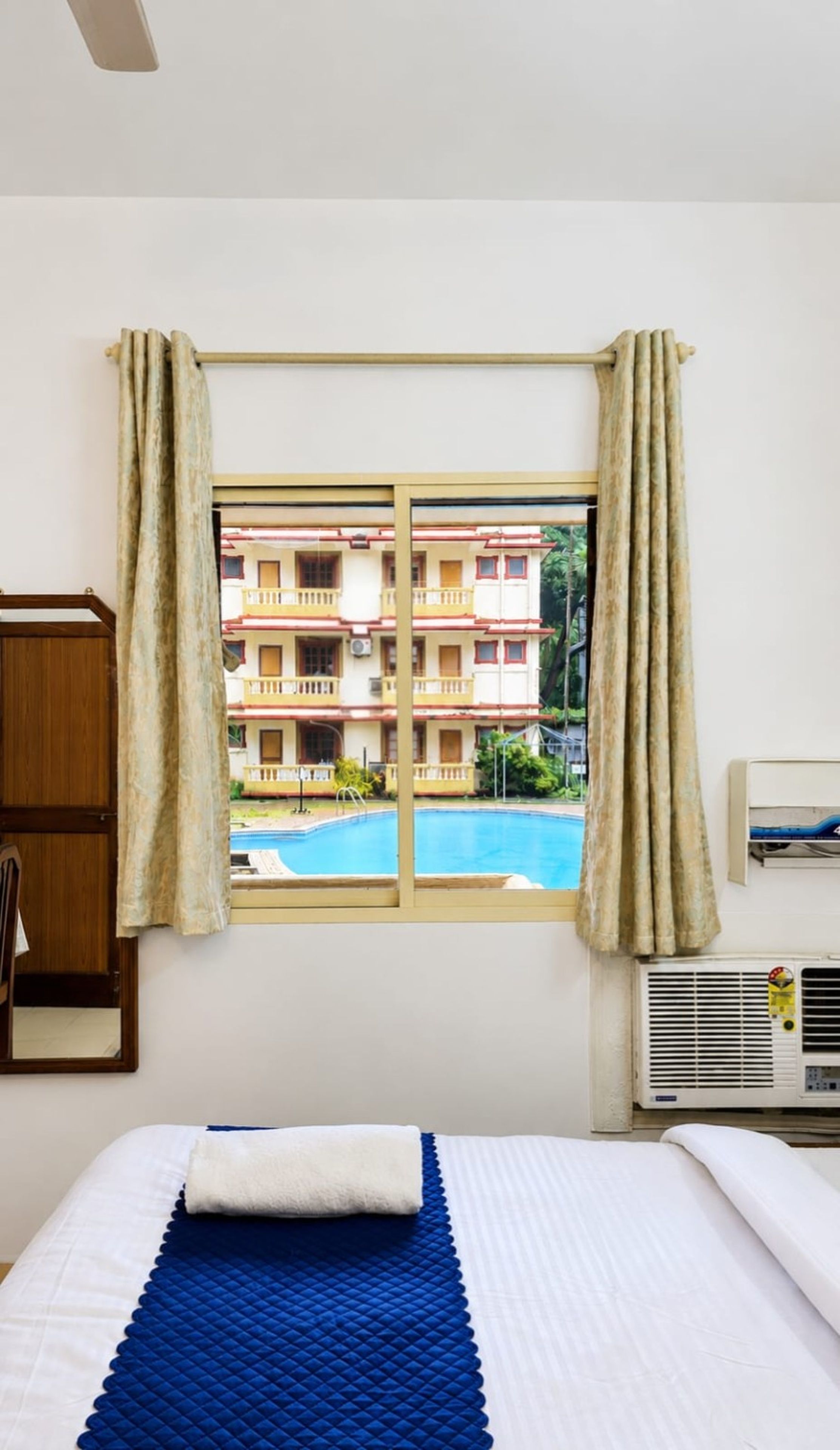 Ein Hotelzimmer mit einem Bett, einem Fenster mit Vorhängen, einer Klimaanlage und Blick auf ein Gebäude mit Balkonen.