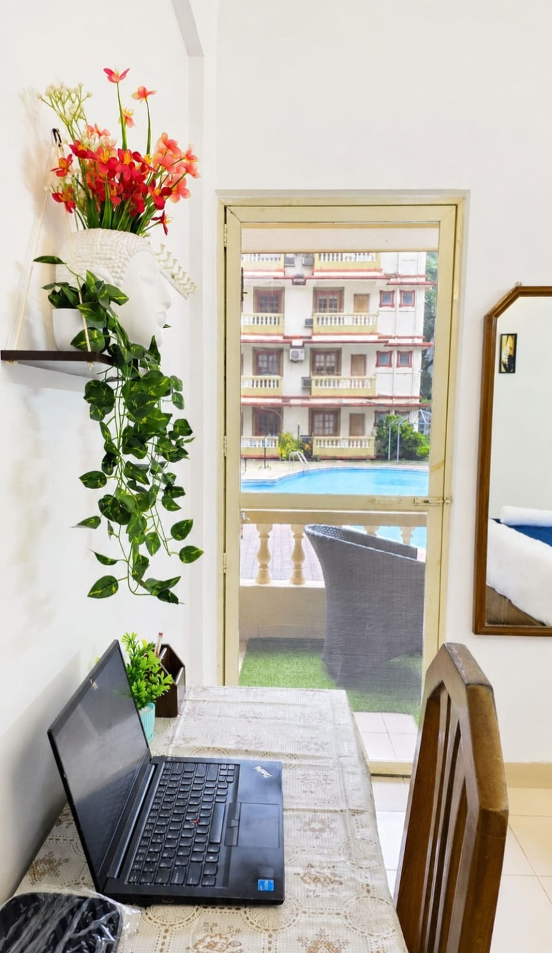 Una habitación con una laptop sobre una mesa, una planta en maceta, un florero con flores y vistas a un edificio con balcones y piscina.