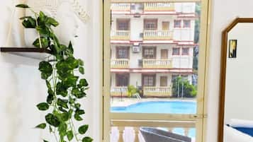 Una habitación con una laptop sobre una mesa, una planta en maceta, un florero con flores y vistas a un edificio con balcones y piscina.