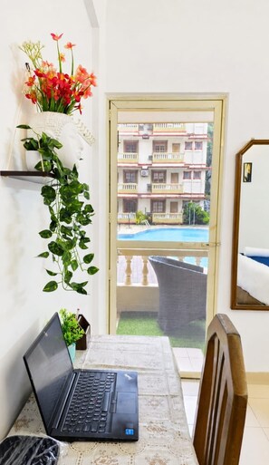 Een kamer met een laptop op een tafel, een kamerplant, een vaas met bloemen en uitzicht op een gebouw met balkons en een zwembad.