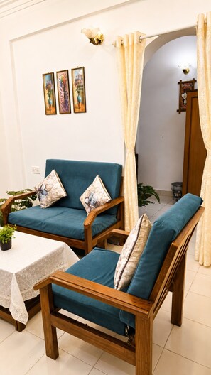 Een kamer met twee houten stoelen met blauwe kussens, een kleine tafel met een witte tafelkleed, en een wand met drie ingelijste foto's.
