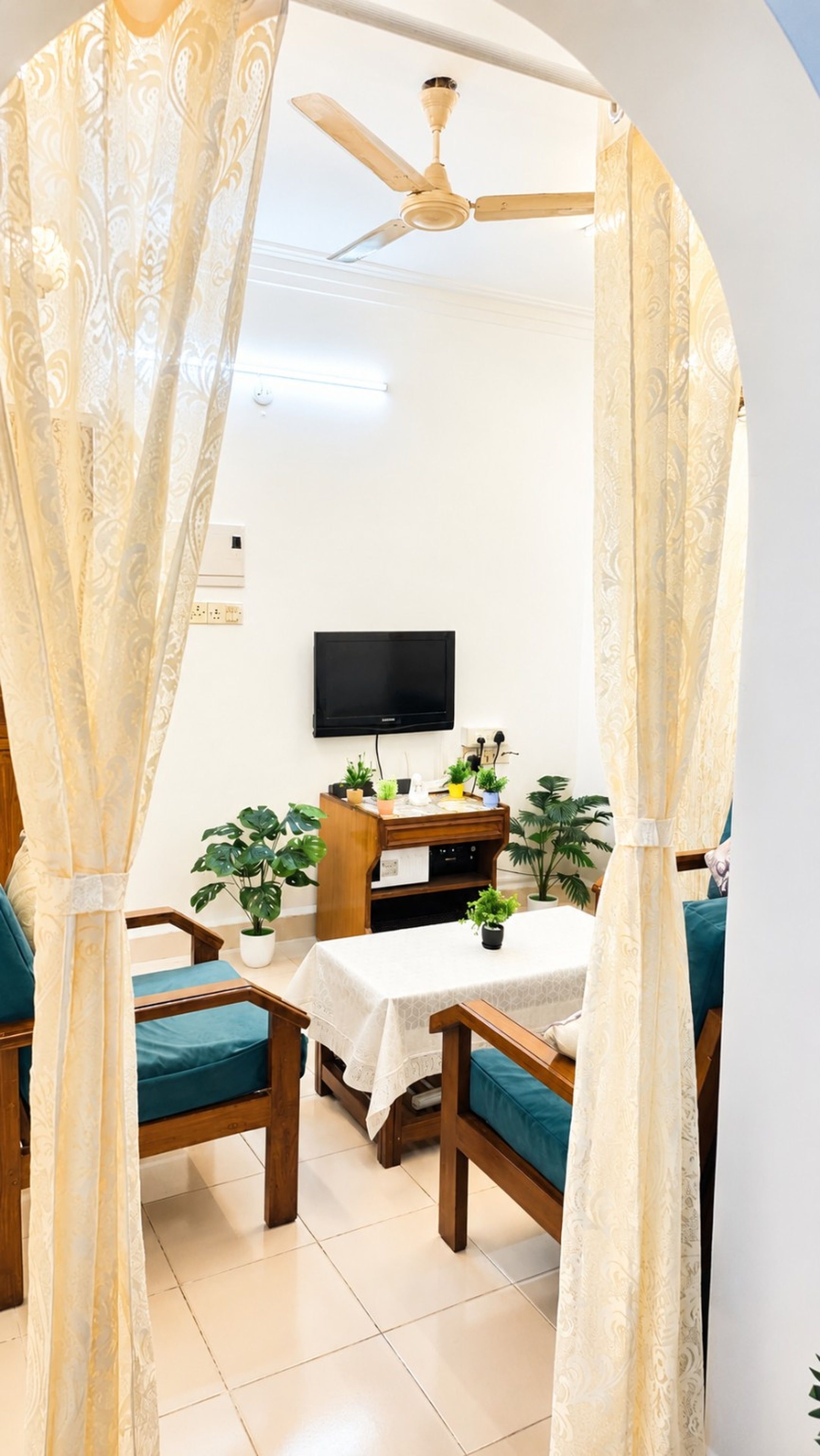 Quarto com ventilador de teto, TV de tela plana na parede, mesa de madeira com toalha branca e duas cadeiras estofadas verdes.