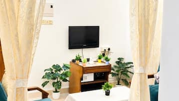 Una habitación con ventilador de techo, televisor de pantalla plana montado en la pared, una mesa de madera con mantel blanco y dos sillas con cojines verdes.