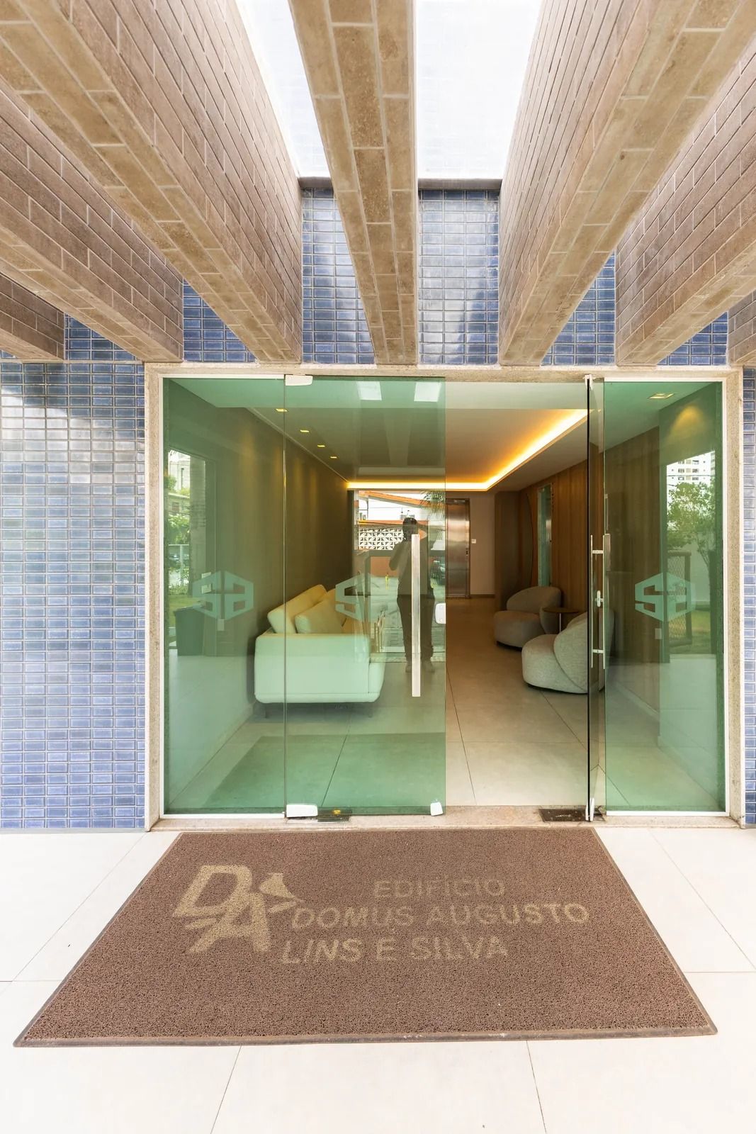 Entrada de um edifício moderno com portas de vidro, área de recepção e elevador visível.