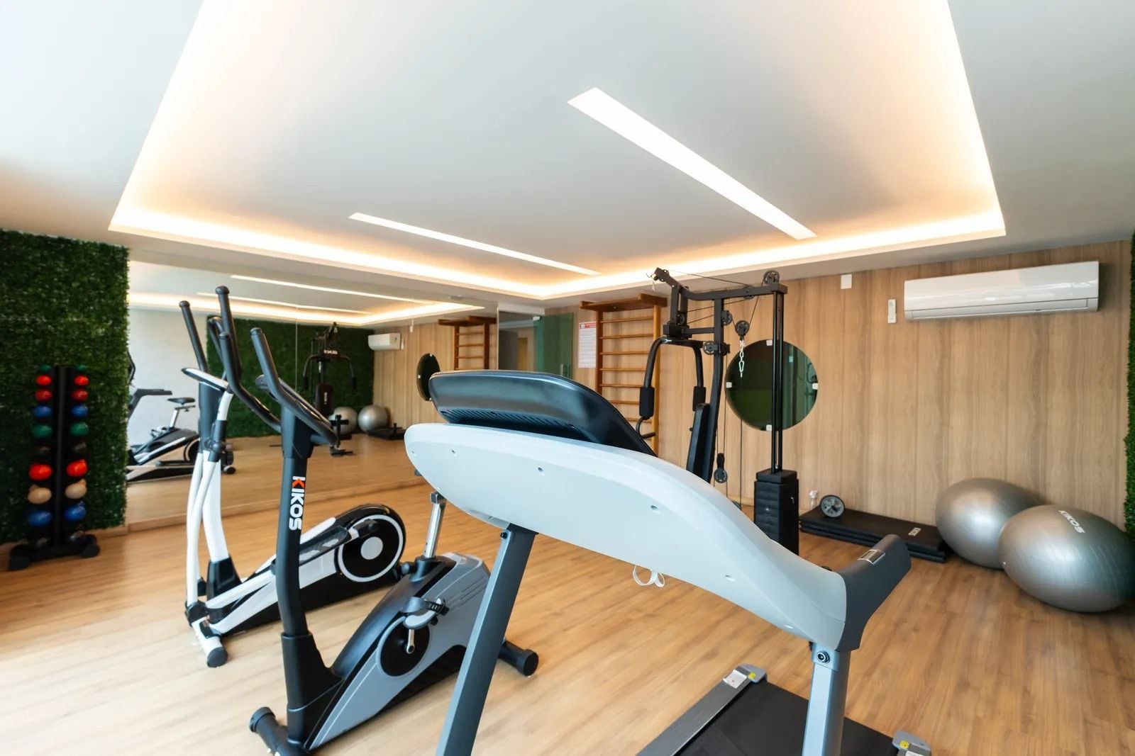 Academia com equipamentos de exercício, parede verde e piso de madeira.