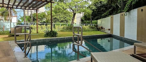 A swimming pool with metal railings, a covered seating area, and a garden with grass and plants.