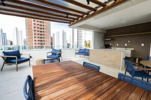 Una terraza en la azotea con mesa de madera, sillas azules, zona de bar y vistas a la ciudad.