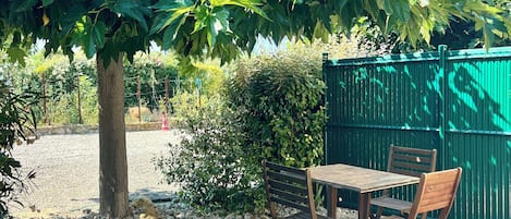 A gravel patio with a wooden table and chairs, surrounded by greenery and a green fence.