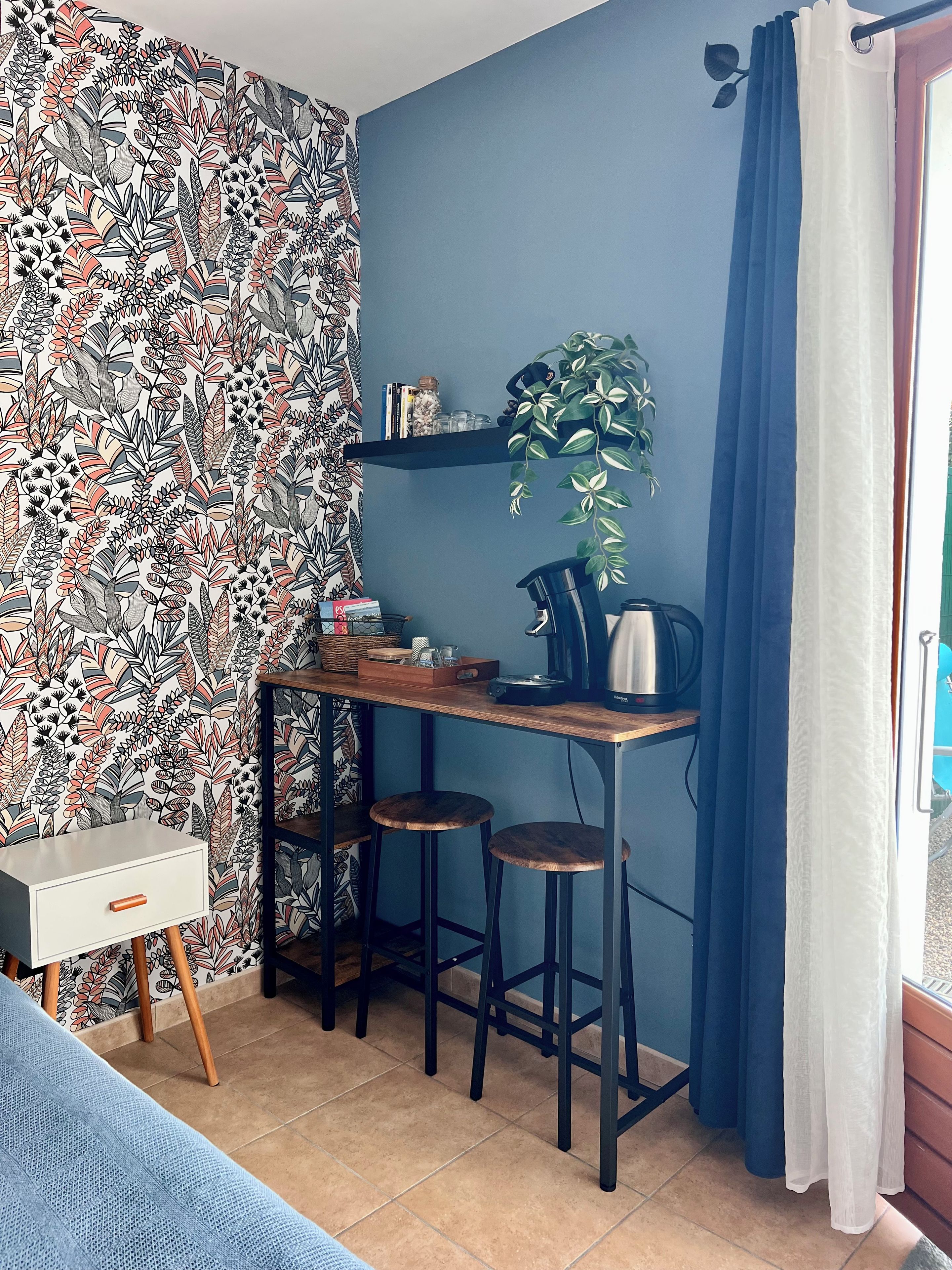 Een kleine, knusse kamer met behang met een patroon, een bureau met een waterkoker, en een potplant.