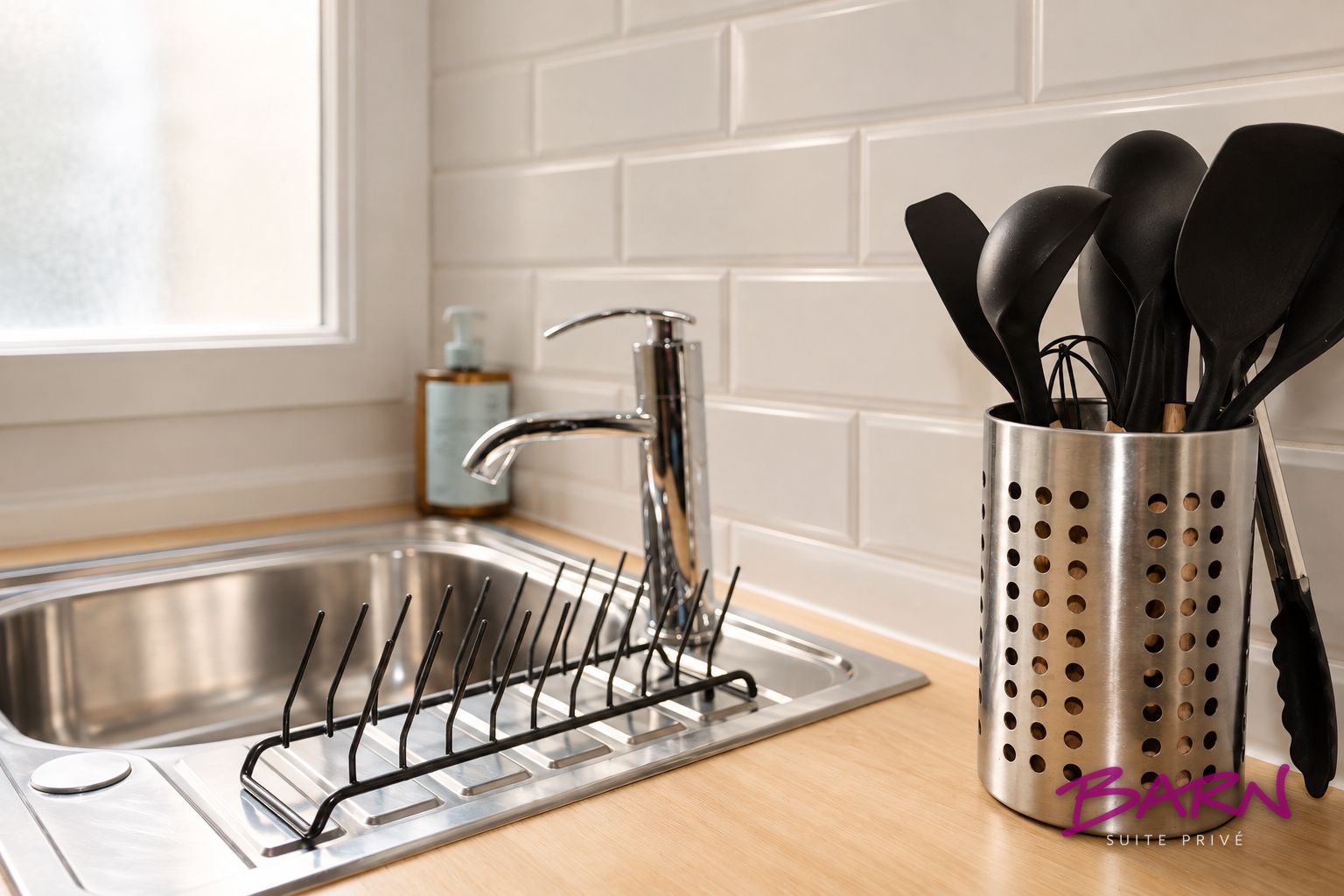 Una cocina con un lavamanos de acero inoxidable, un portalaputazos con utensilios negros y una botella de jabón para manos en la encimera.