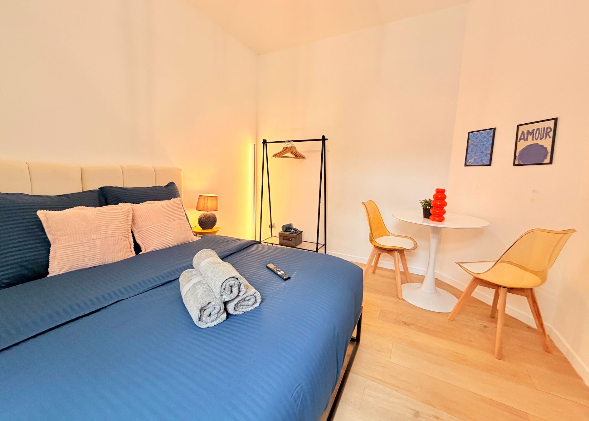 A hotel room with a bed, a bedside table with a lamp, a chair, a small table with a potted plant, and two framed pictures on the wall.