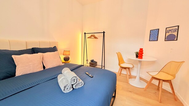 A hotel room with a bed, a bedside table with a lamp, a chair, a small table with a potted plant, and two framed pictures on the wall. - Le BARN -> a stylish urban retreat ❤️ (Le Havre)