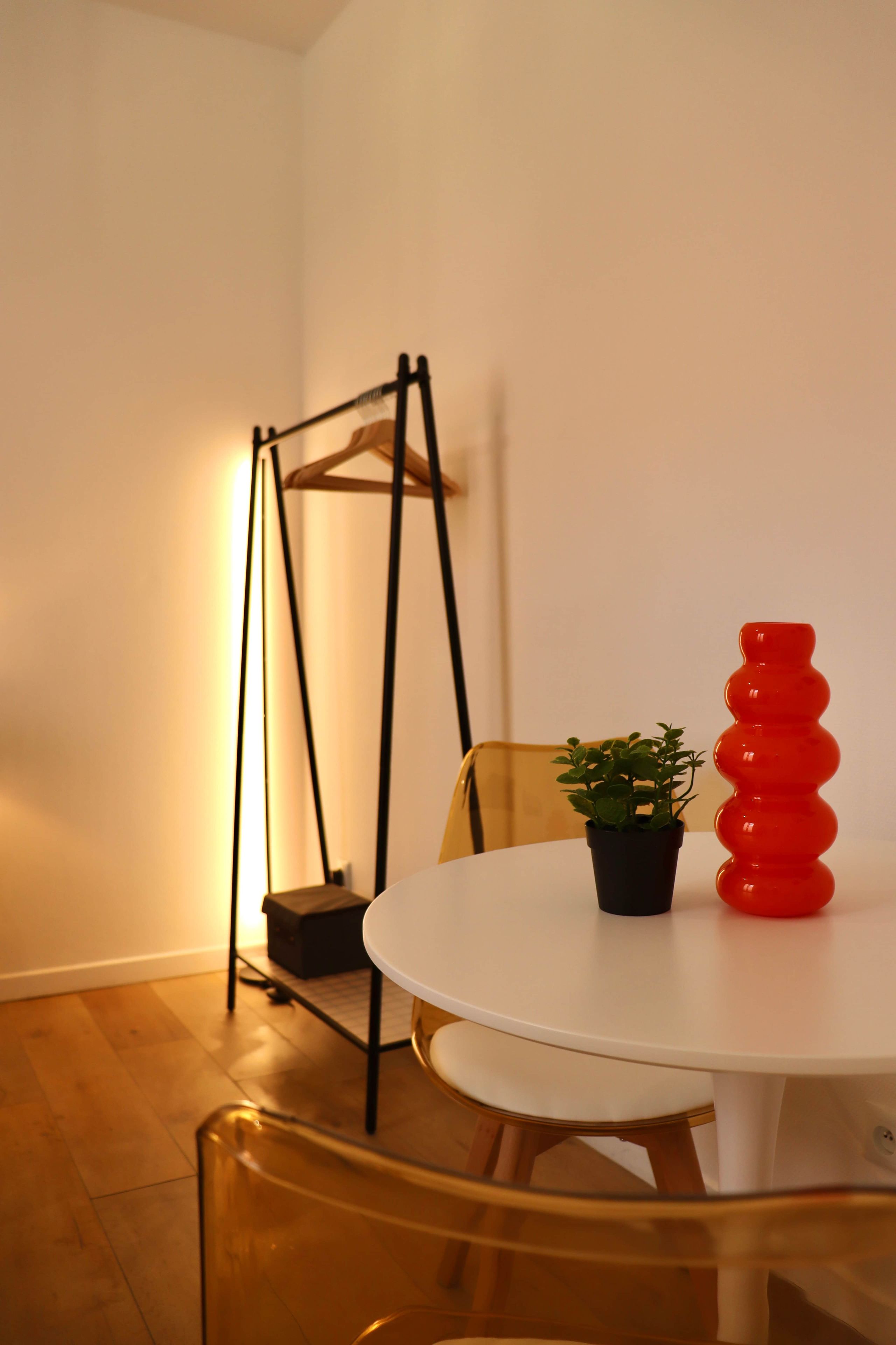 A room with a white table, a black hanging chair, a small potted plant, and a red vase.